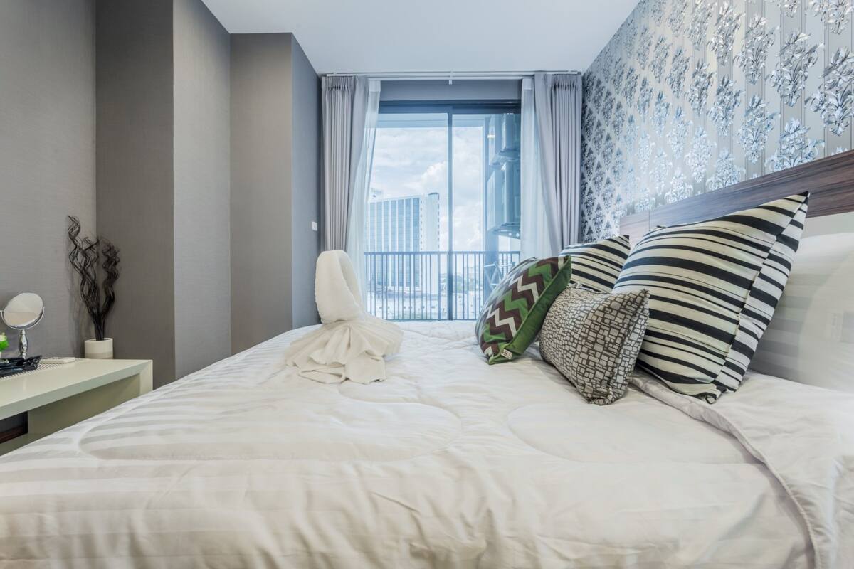 A modern hotel room with a large bed, a bedside table with a mirror, and a view of the city through a large window.