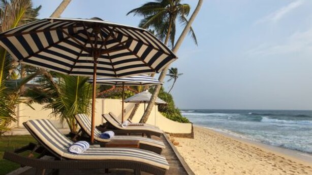 A beachside lounge area with striped lounge chairs and a large umbrella, palm trees, and the ocean in the background. - Isle Of Thalpe (Unawatuna)
