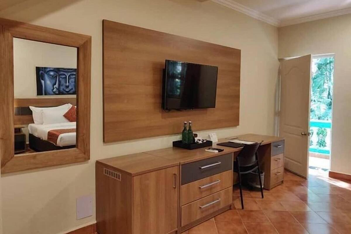 A hotel room with a bed, a wooden cabinet, a desk with a chair, a television mounted on the wall, and a large mirror.