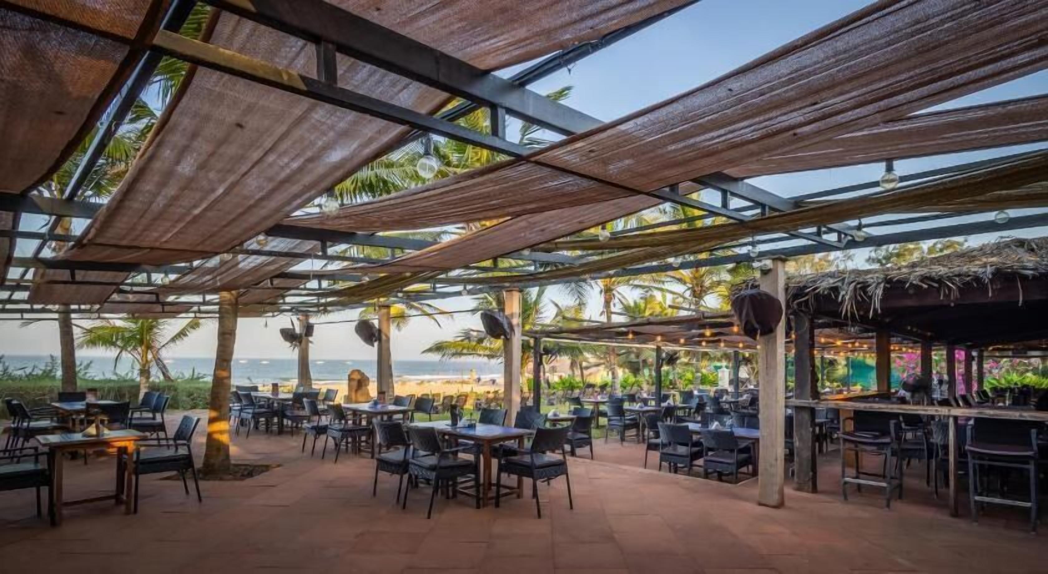 An outdoor restaurant with tables and chairs, a bar area, and a view of the beach and ocean.