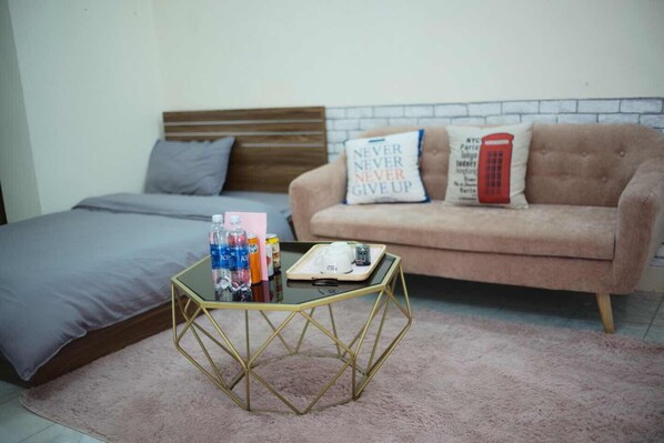 A bedroom with a bed, a glass coffee table, a pink rug, a sofa with two decorative pillows, and a wall with a brick pattern.