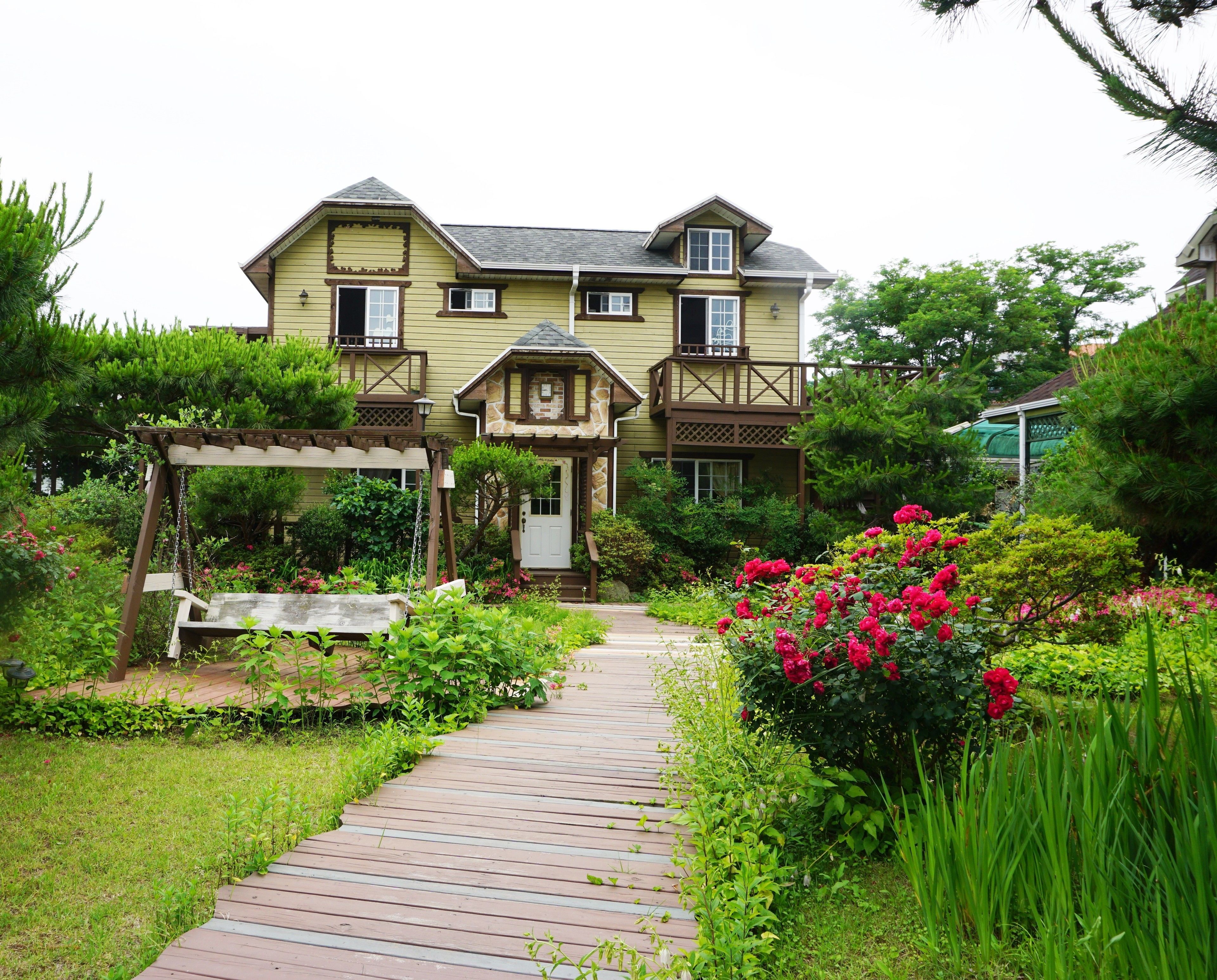 A two-story house with a wooden deck leading to the front door, surrounded by lush greenery and flowering plants.