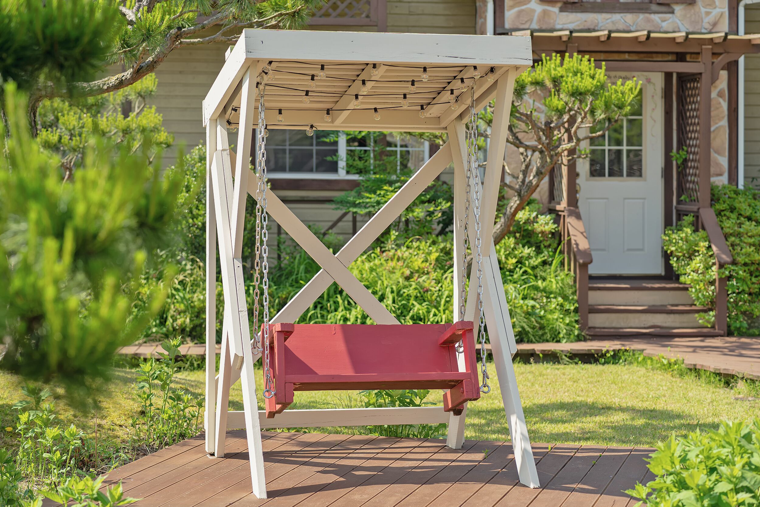 A white swing set with a red seat in a garden area.