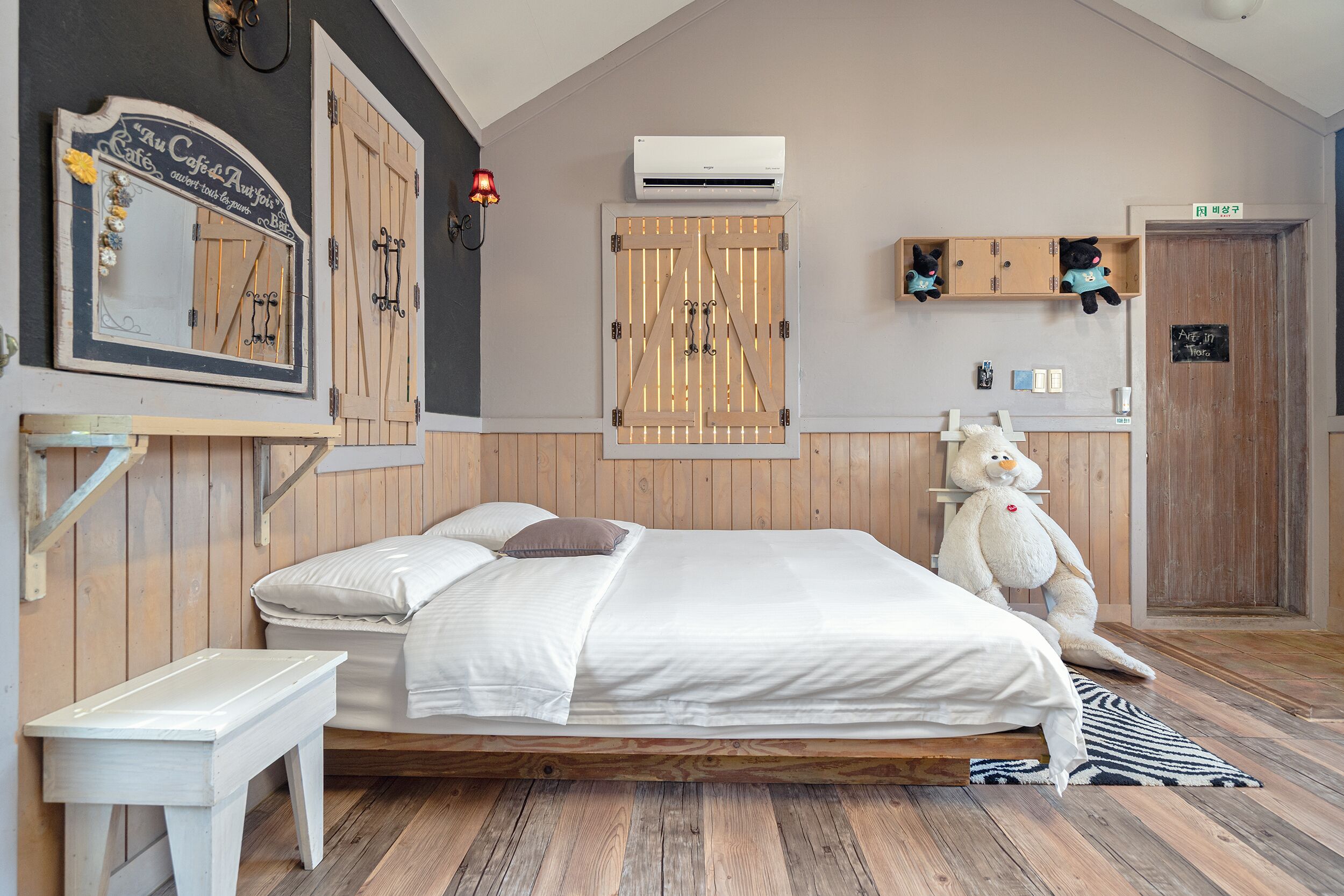 A bedroom with a wooden bed, a white bedside table, a large teddy bear, a mirror, and a wardrobe with a wooden door.
