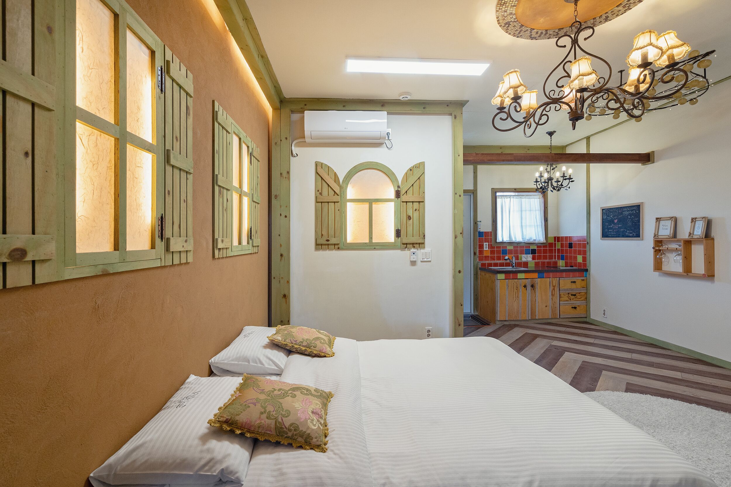 A hotel room with a large bed, two pillows, a chandelier, an air conditioning unit, and a view of the kitchen.