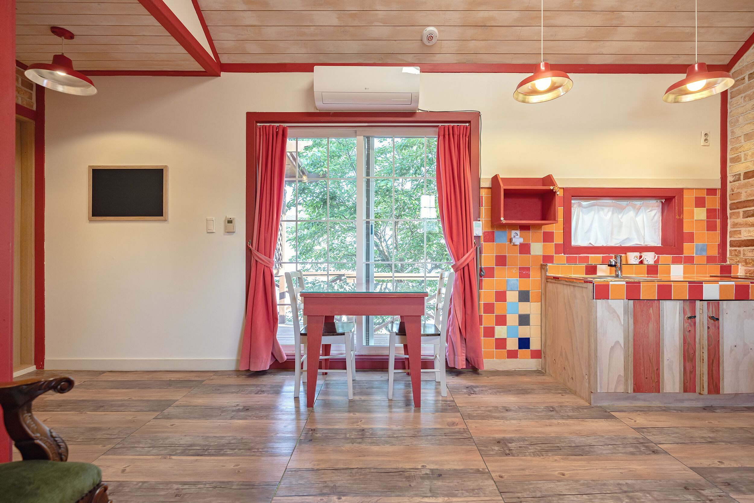 Une pièce avec un plancher en bois, une table entourée de chaises, une fenêtre avec des rideaux rouges, un coin cuisine avec un pare-feu coloré et un climatiseur fixé au mur.