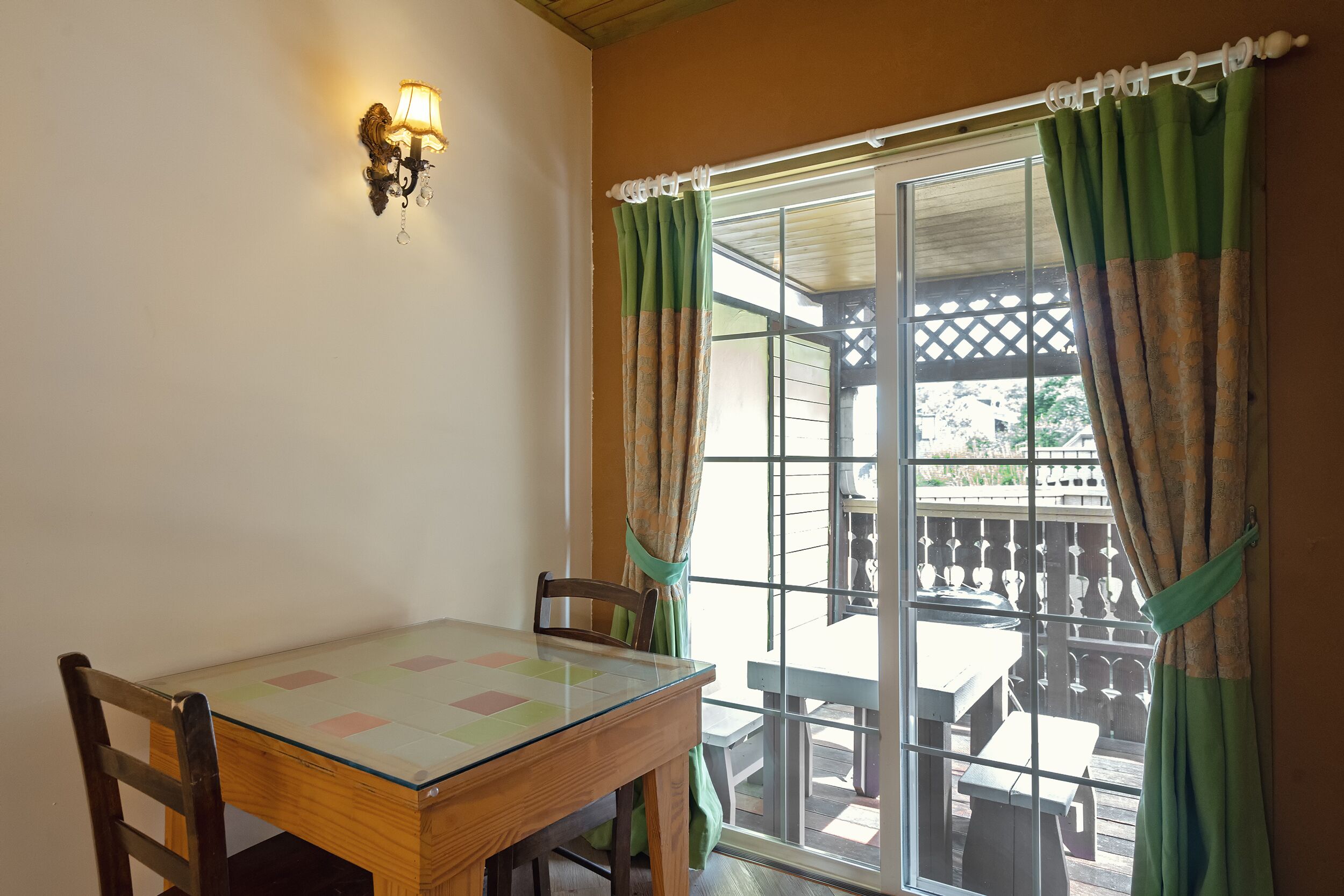 Uma área de refeições com mesa de vidro, cadeiras de madeira, luminária fixada na parede e vista para um pátio externo através de uma porta de vidro deslizante com cortinas verdes.
