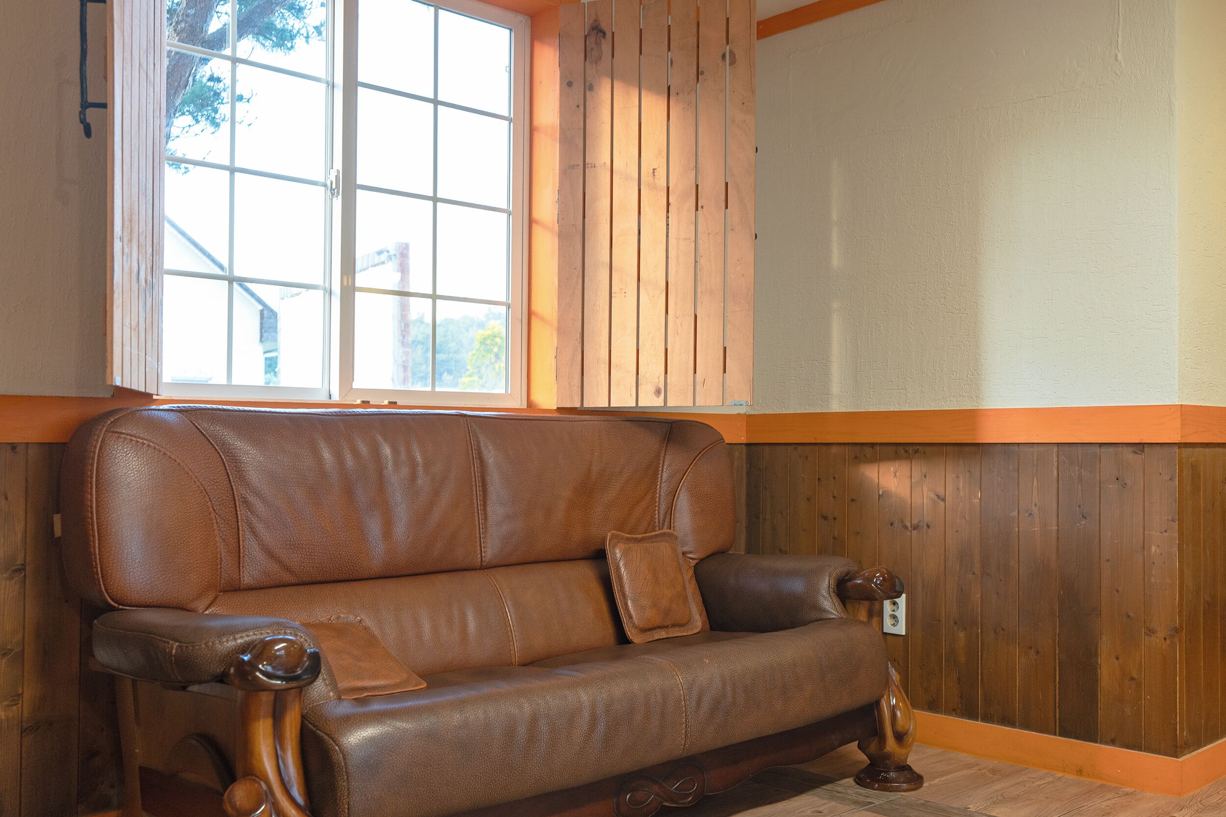 A brown leather sofa with wooden legs and a cushion, positioned against a wooden wall with a window above it.