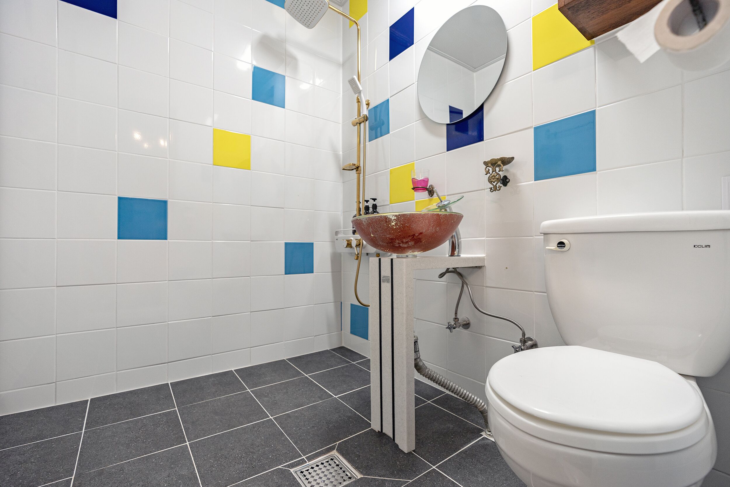 A bathroom with a white toilet, a sink with a red basin, and a shower. The walls are tiled with a pattern of white, blue, and yellow squares.