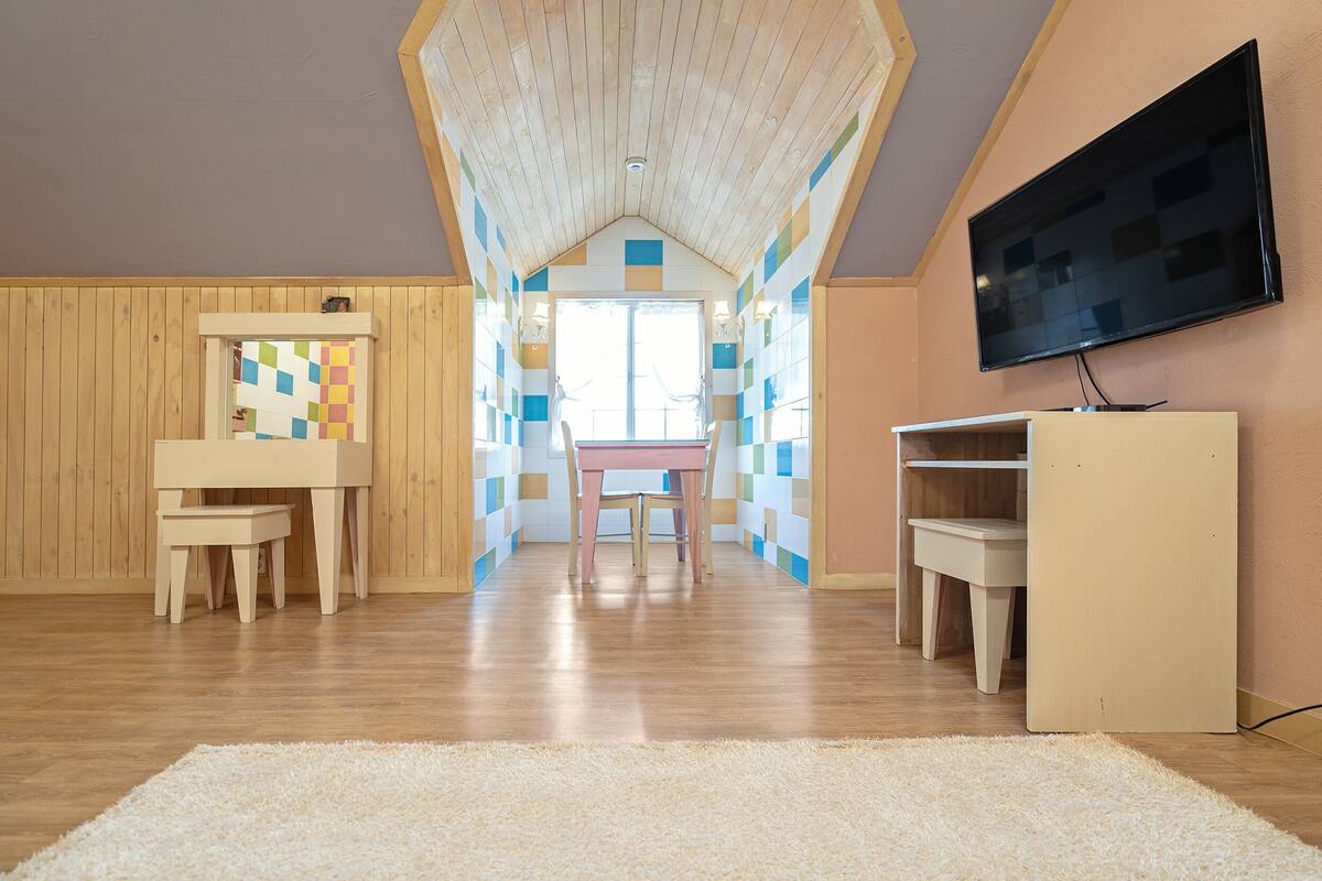 A room with a wooden ceiling, a TV mounted on the wall, a small table with chairs, and a colorful checkered wall.