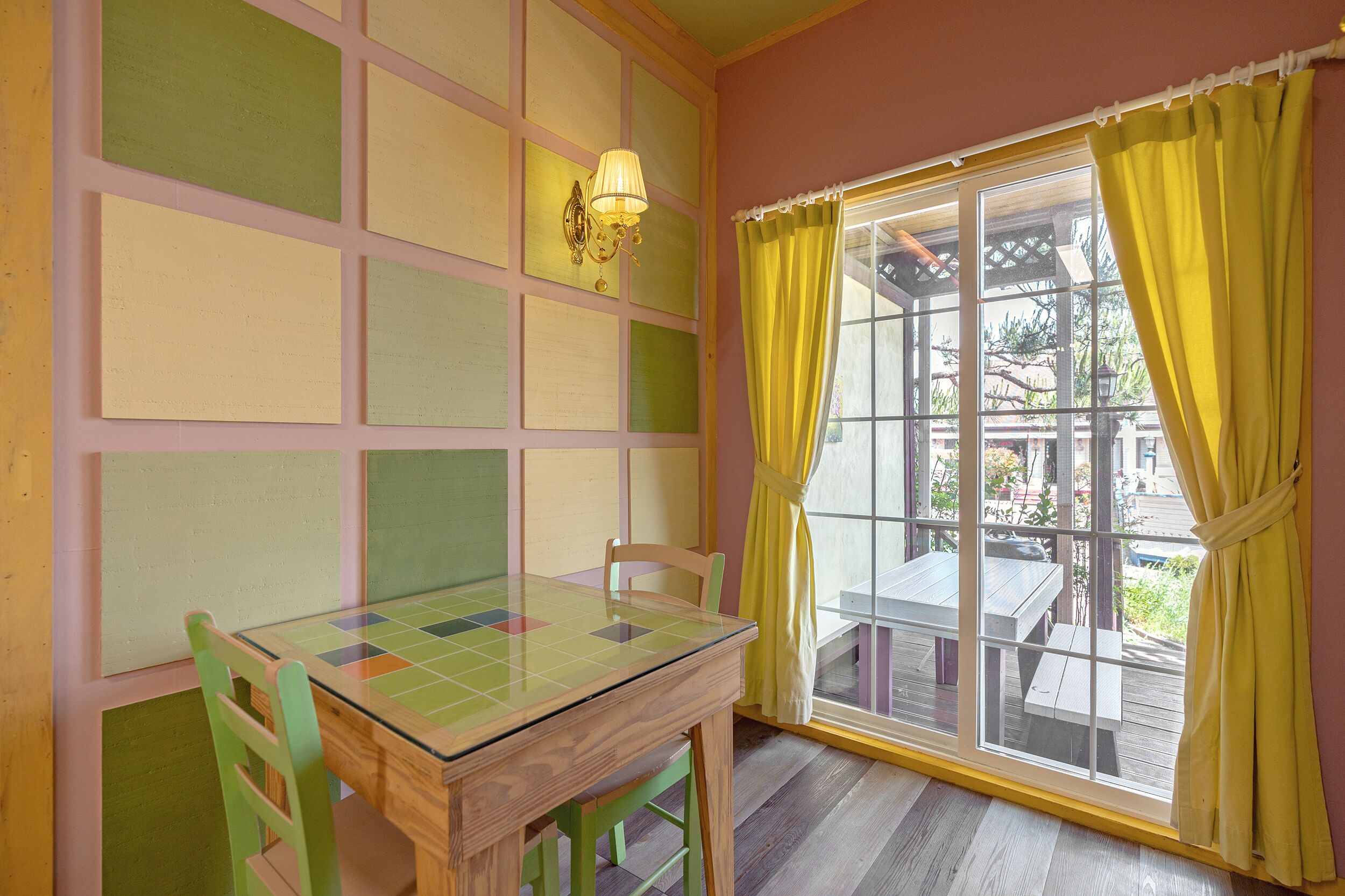 A room with a glass-top table and chairs, a window with yellow curtains, and a wall with a grid of pastel-colored panels.
