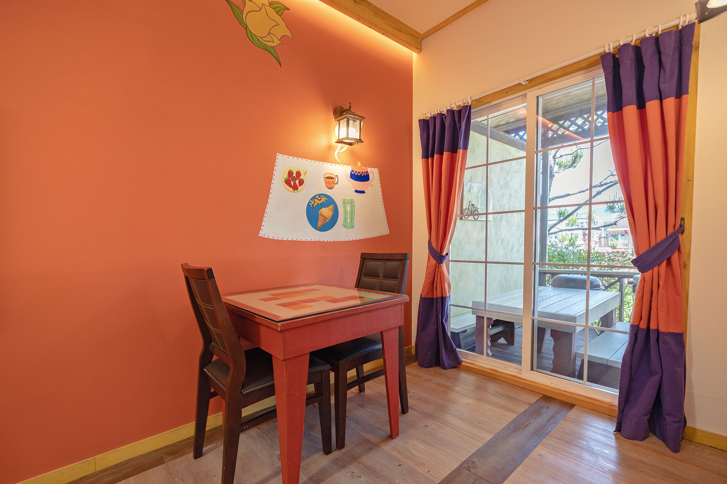 A dining area with a wooden table and chairs, a wall-mounted light, and a window with purple curtains.