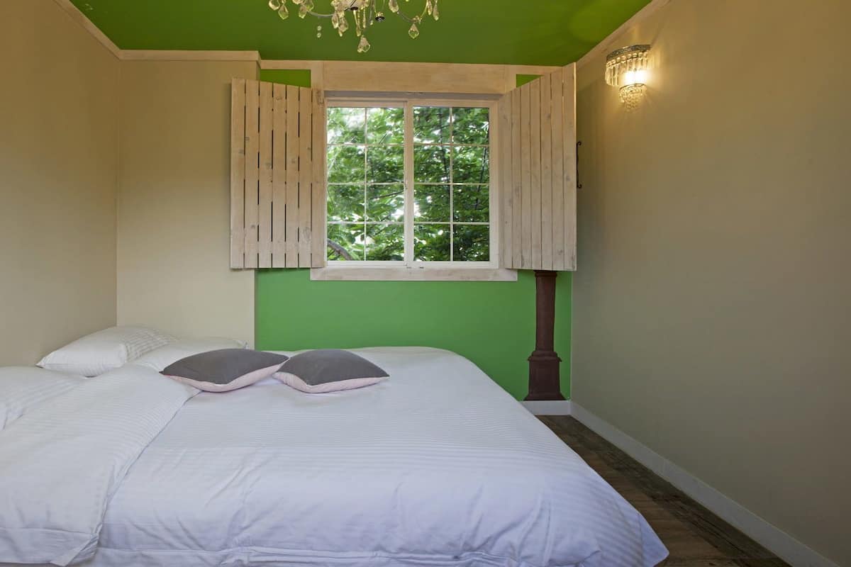 A bedroom with a bed, a window with shutters, a chandelier, and a wall-mounted light fixture.