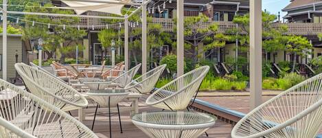 A covered outdoor seating area with wicker chairs and tables, surrounded by greenery and residential buildings.