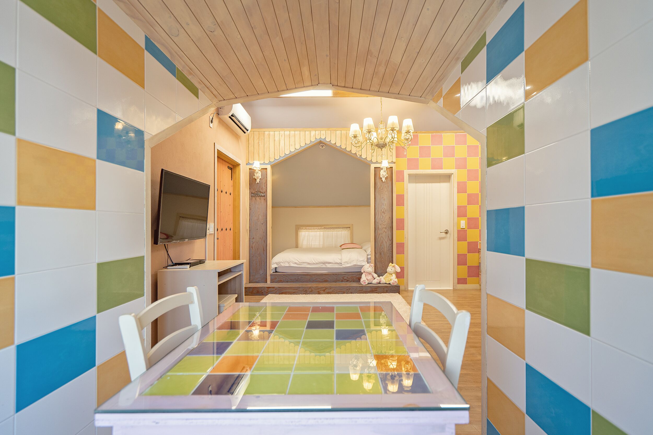 Um quarto com piso xadrez, cama com cabeceira branca, televisão sobre suporte e um candelabro.