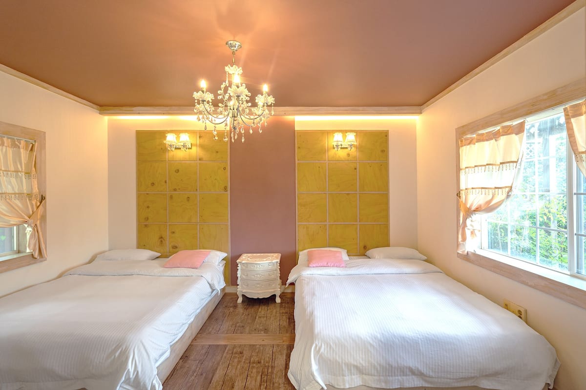 A room with two beds, a chandelier, wooden paneling on the walls, and a window with curtains.