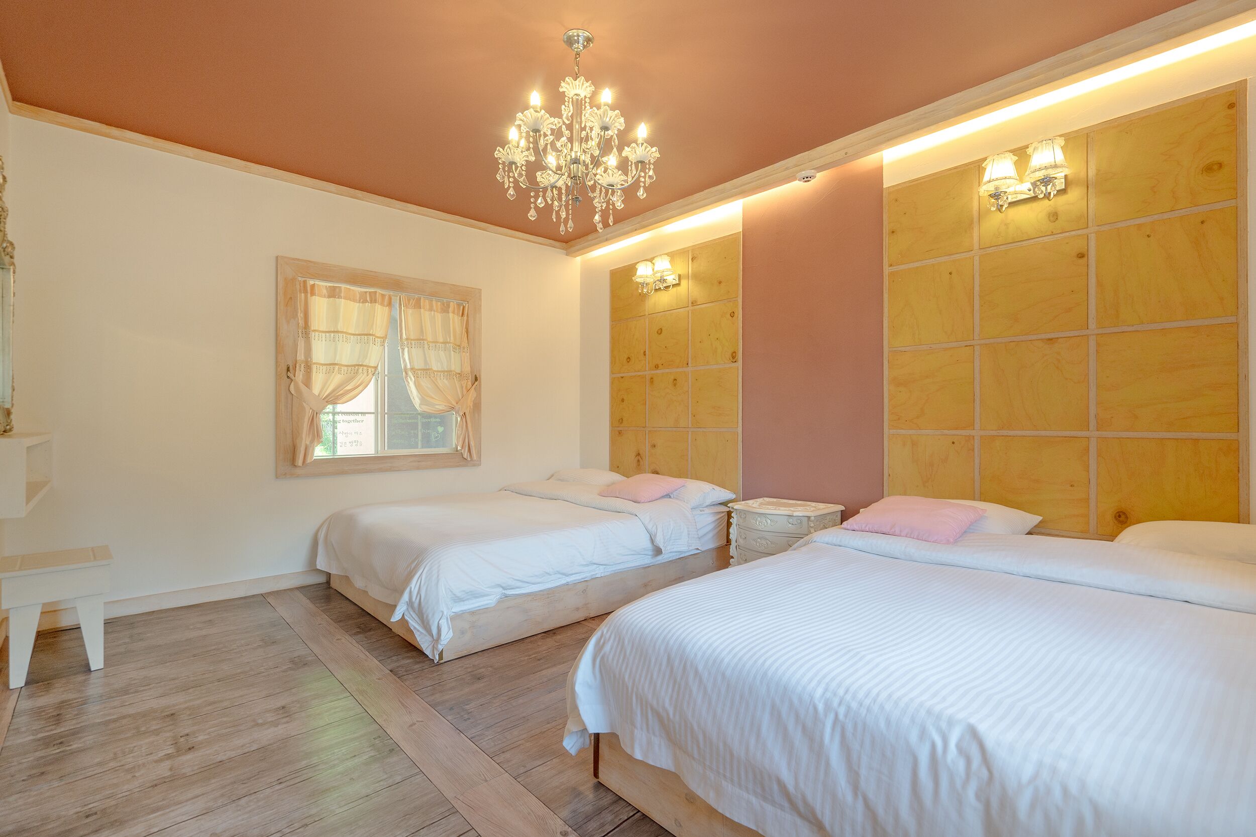 Une chambre avec deux lits, un lustre, une fenêtre avec des rideaux et une tête de lit en bois.