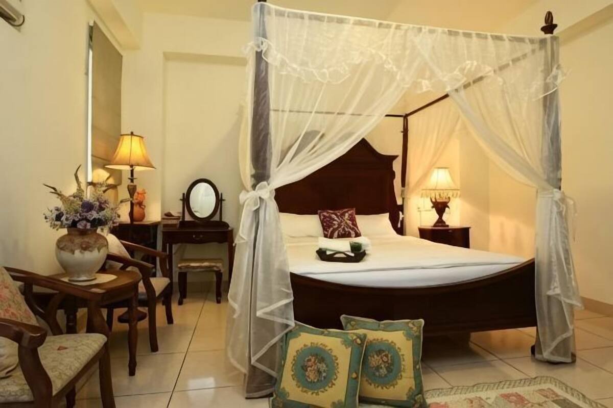 A four-poster bed with a canopy, a bedside table with a lamp, a chair, and a small table with a vase of flowers.