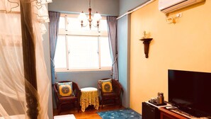 A living room with two wicker chairs, a small round table, a blue patterned rug, a television on a wooden cabinet, and a window with curtains. - Hope Sea Bed and Breakfast (Hualien City)