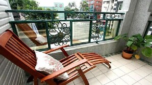 A wooden bench with a cushion on a balcony with a metal railing, potted plants, and a view of buildings in the background. - Hope Sea Bed and Breakfast (Hualien City)