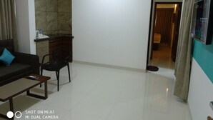 A living room with a dark sofa, a wooden coffee table, a black chair, a kitchen area with a counter and cabinets, and a hallway leading to another room.