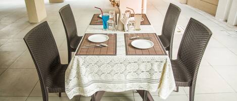 A table set for four with a white lace tablecloth, dark wicker chairs, and a vase with flowers.