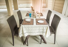 A table set for four with a white lace tablecloth, dark wicker chairs, and a vase with flowers.