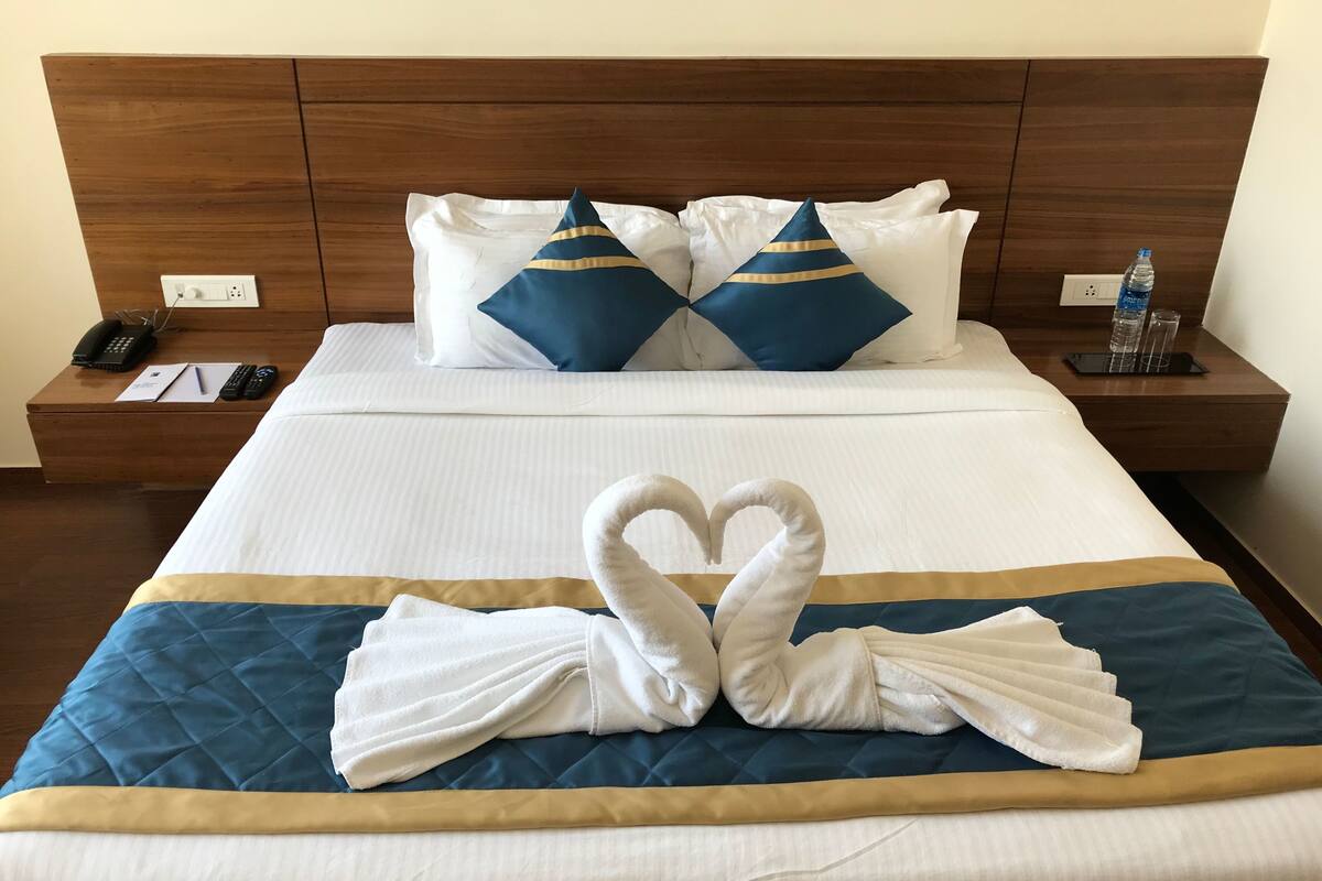 A neatly made bed with a heart-shaped towel arrangement, a wooden headboard, and a bedside table with a phone, remote controls, and a water bottle.