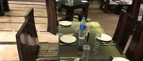 A dining area with glass tables, wooden chairs, and a buffet counter in the background.