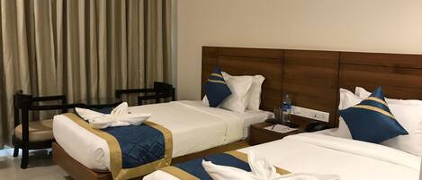 A hotel room with two beds, each with a blue and gold bedspread, white pillows, and a neatly folded towel on each bed. There is a wooden nightstand with a phone and a bottle of water.