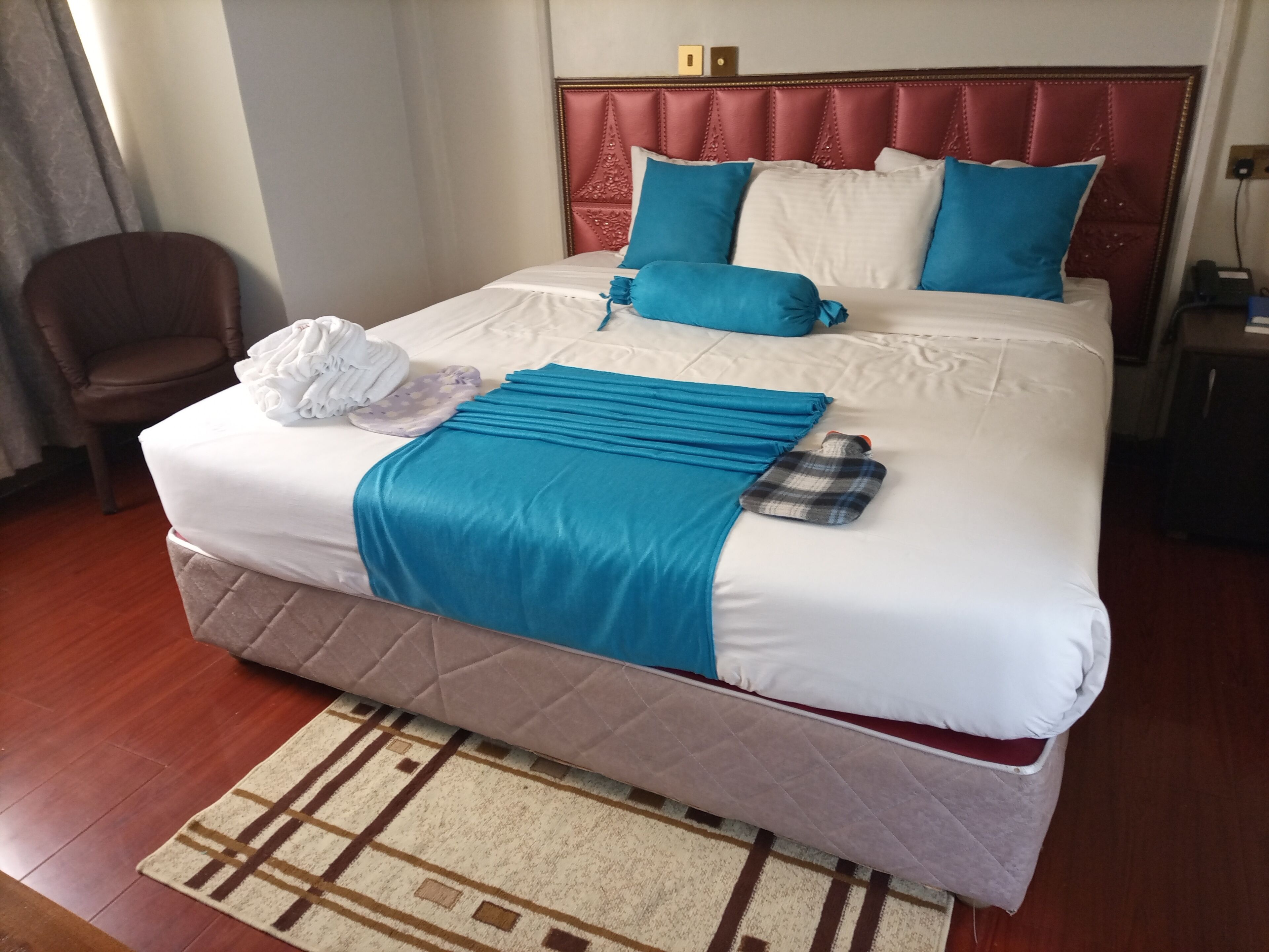 Una cama bien tendida con una colcha azul y almohadas, una almohada de cuadros y una toalla doblada al lado.