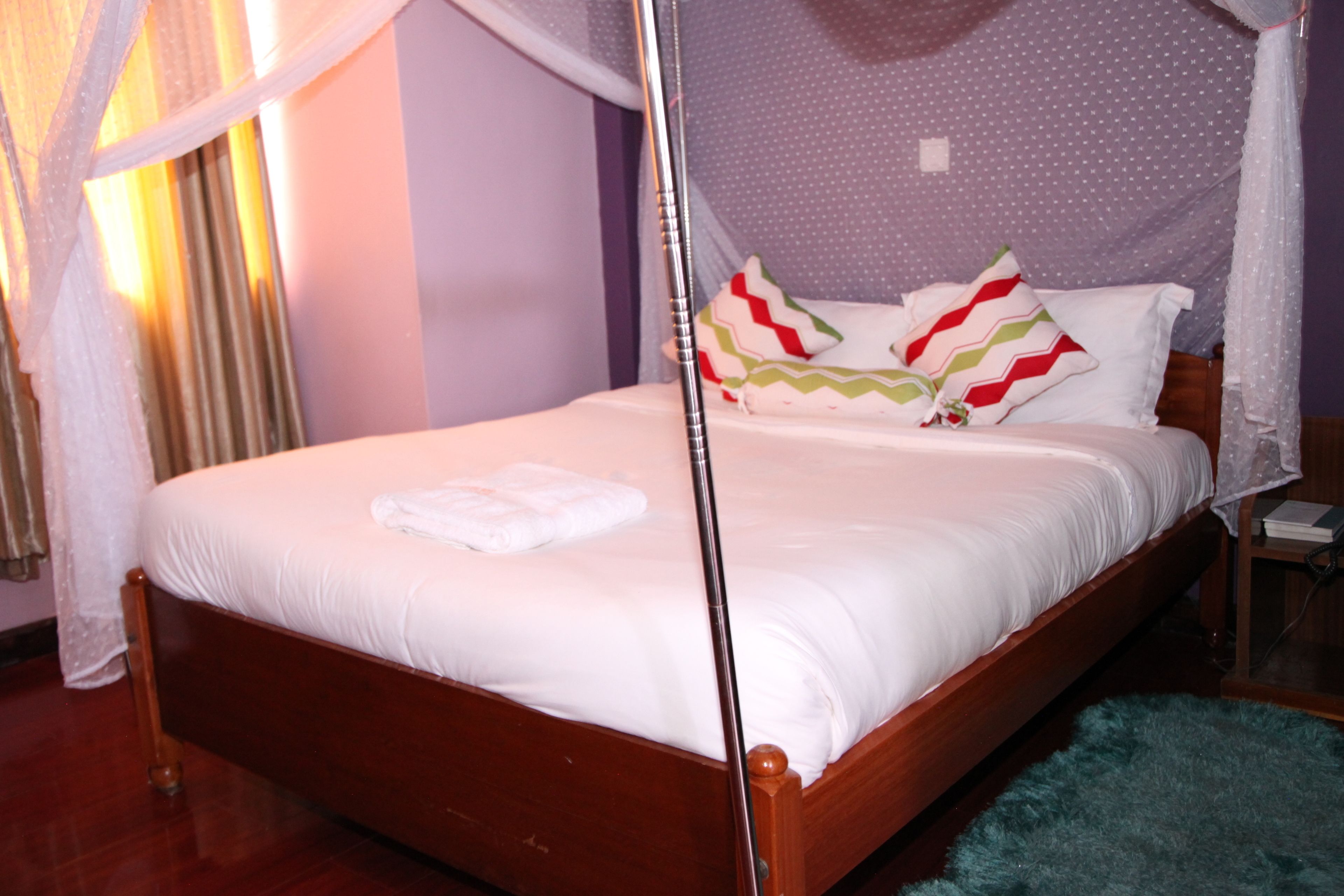 A neatly made bed with white linens and a wooden headboard, a bedside table with a lamp, and a teal rug on the floor.