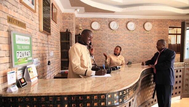 A reception desk with a brick wall, clocks labeled with country names, and a till number sign. - Sunshine Hotel (Kericho)
