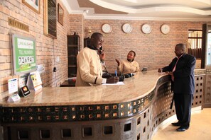 A reception desk with a brick wall, clocks labeled with country names, and a till number sign. - Sunshine Hotel (Kericho)