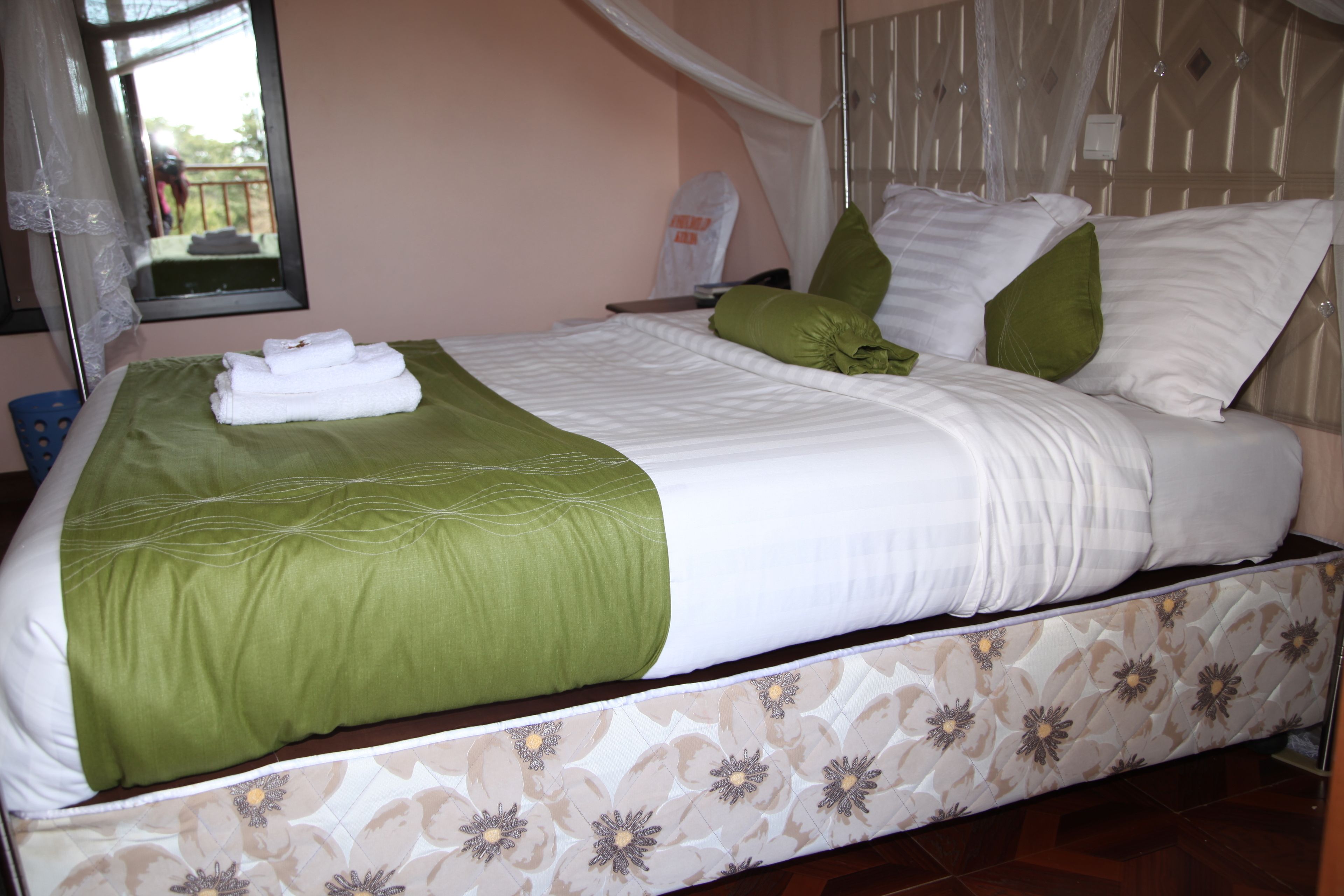 Una cama bien hecha con edredón verde y blanco, cabecera estampada con motivos florales y una ventana con vistas al exterior.