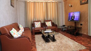 A living room with a brown leather sofa, a glass coffee table, and a television on a stand. - Sunshine Hotel (Kericho)