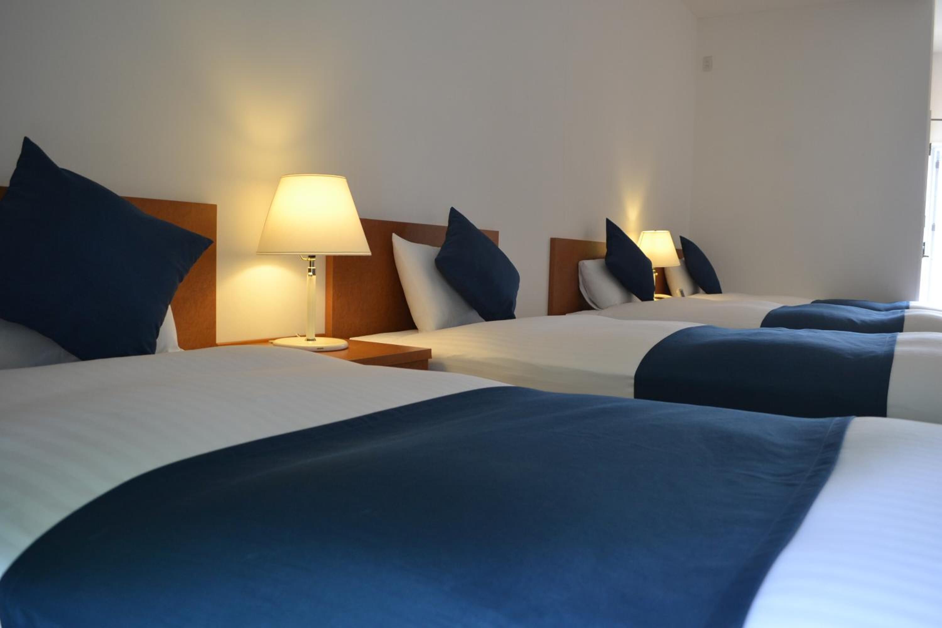 A hotel room with three beds, each with a headboard, bedside lamps, and blue bedding.