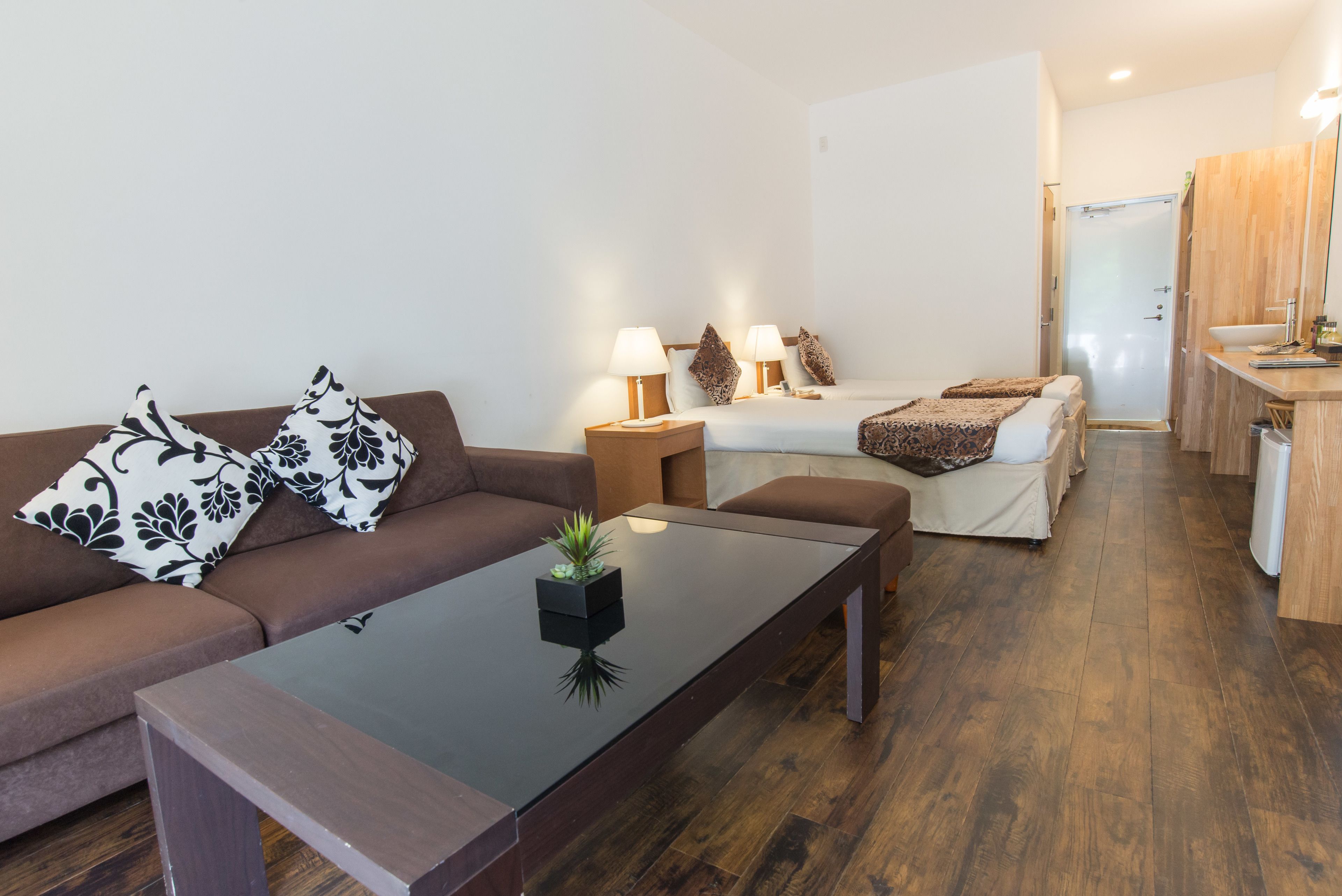 A modern living room with a brown sofa, a glass coffee table, and a white bed with two bedside tables.