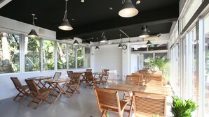 A spacious indoor area with multiple wooden tables and chairs arranged for dining or meetings. Large windows allow natural light and provide views of greenery outside.