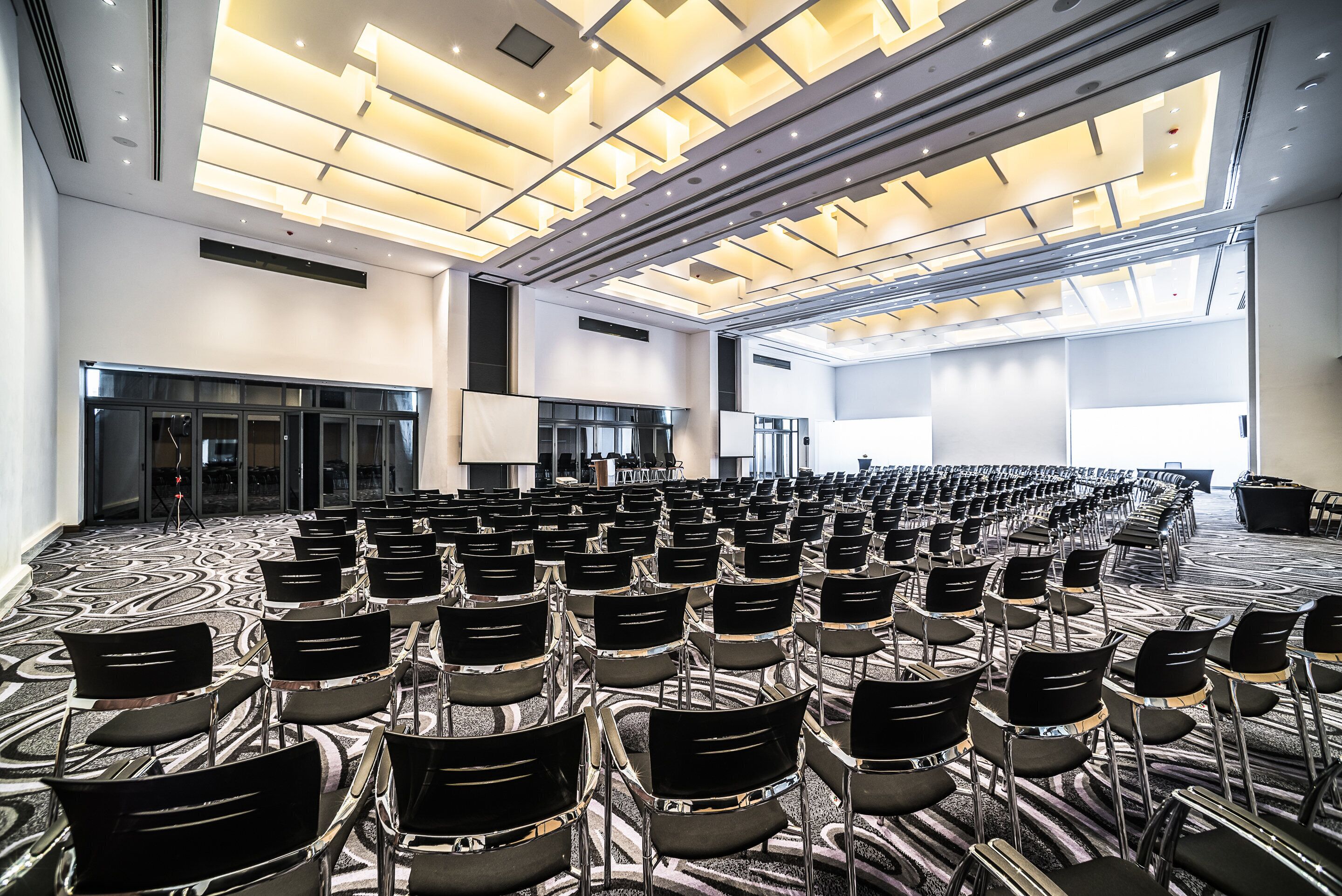Un amplio salón de conferencias con filas de sillas, una alfombra estampada y un diseño de techo moderno.