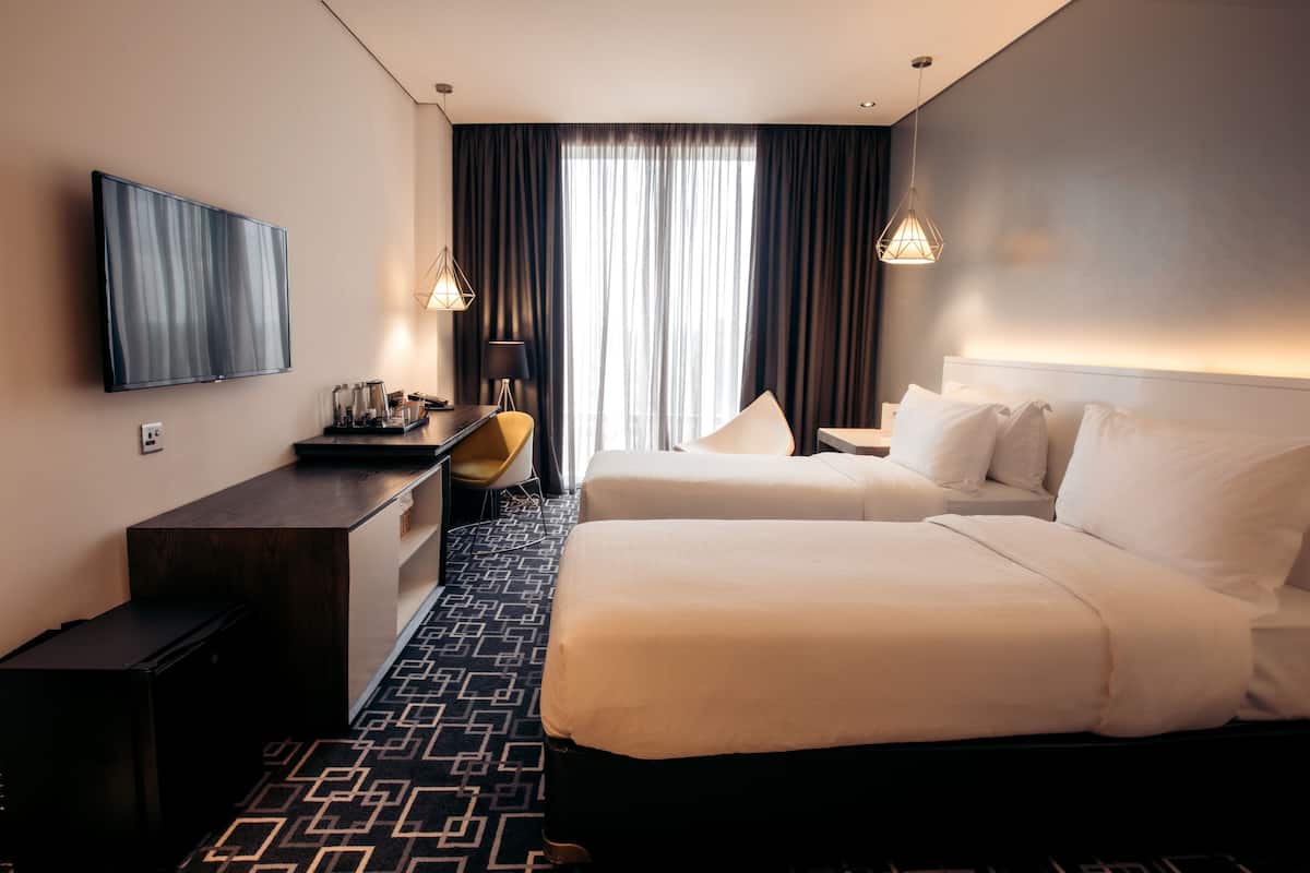 A hotel room with two beds, a desk, a TV, and a large window with curtains.
