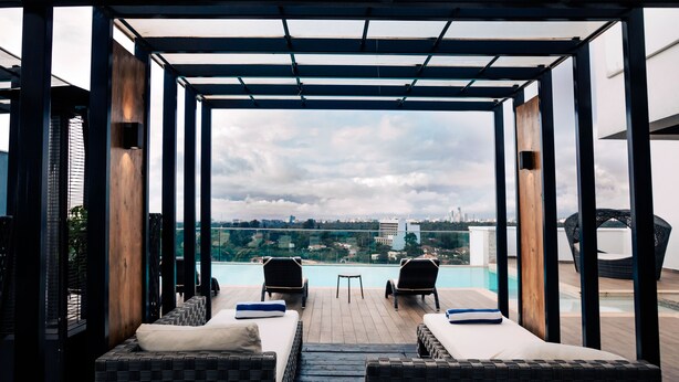 A rooftop terrace with a glass balustrade, two wicker sofas with cushions, a small pool, and a city view. - Trademark Hotel (Nairobi)