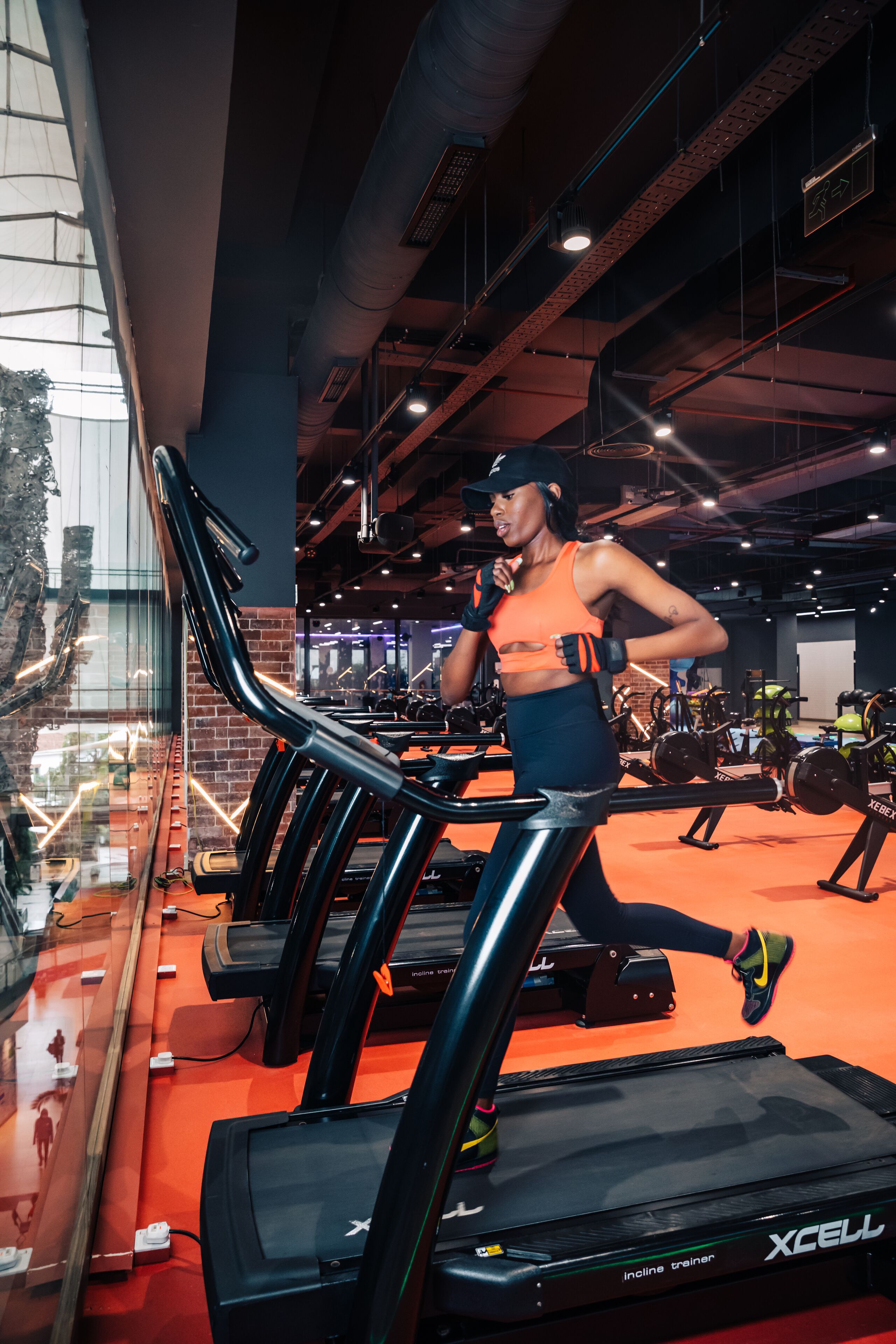 A woman running on a treadmill in a gym with visible exercise equipment and a brick wall in the background.