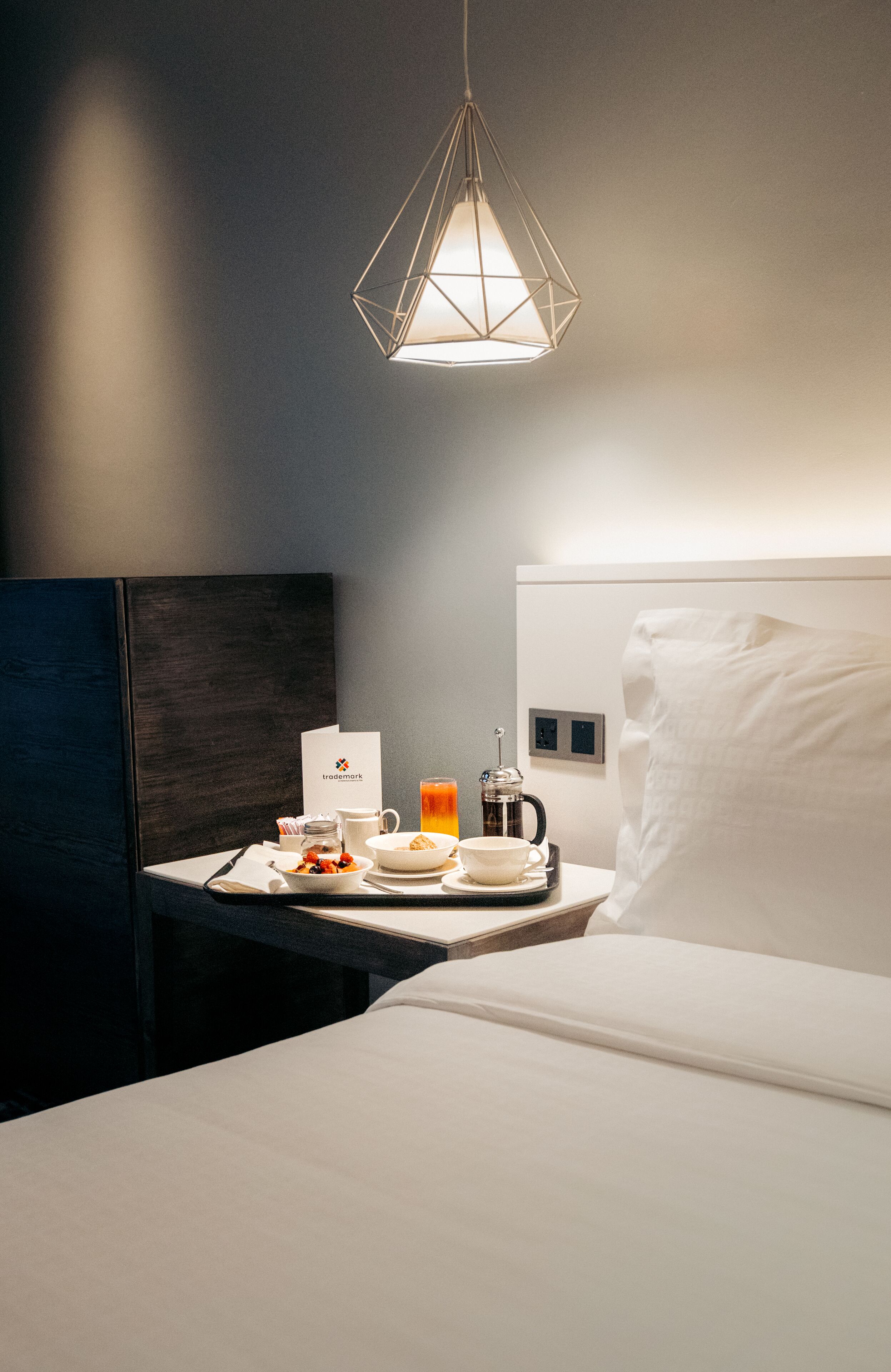 A hotel room with a neatly made bed, a bedside table with a coffee maker, a bowl of fruit, and a white pillow.