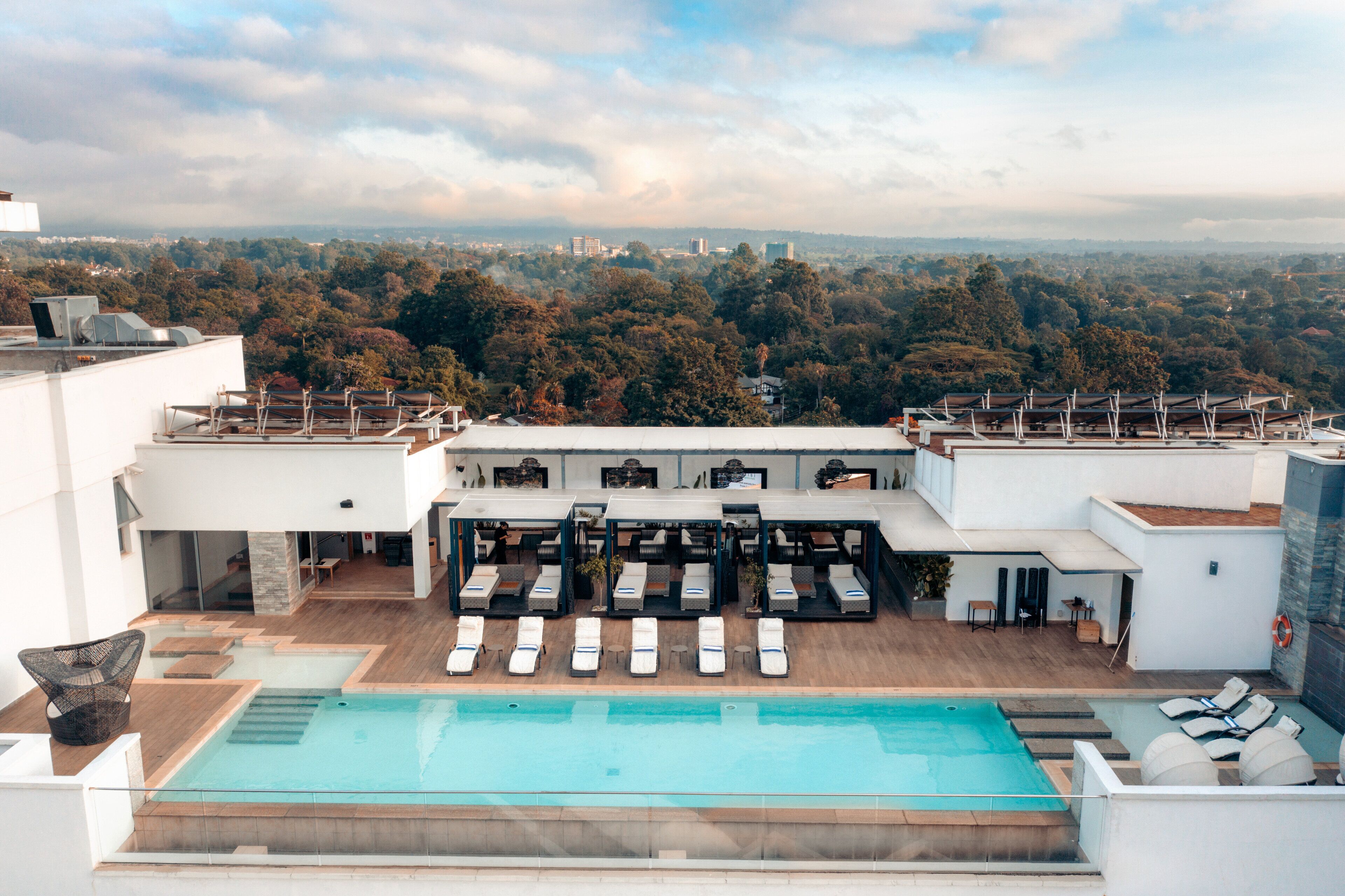 A rooftop pool area with multiple lounge chairs and a pool, surrounded by a cityscape and trees.