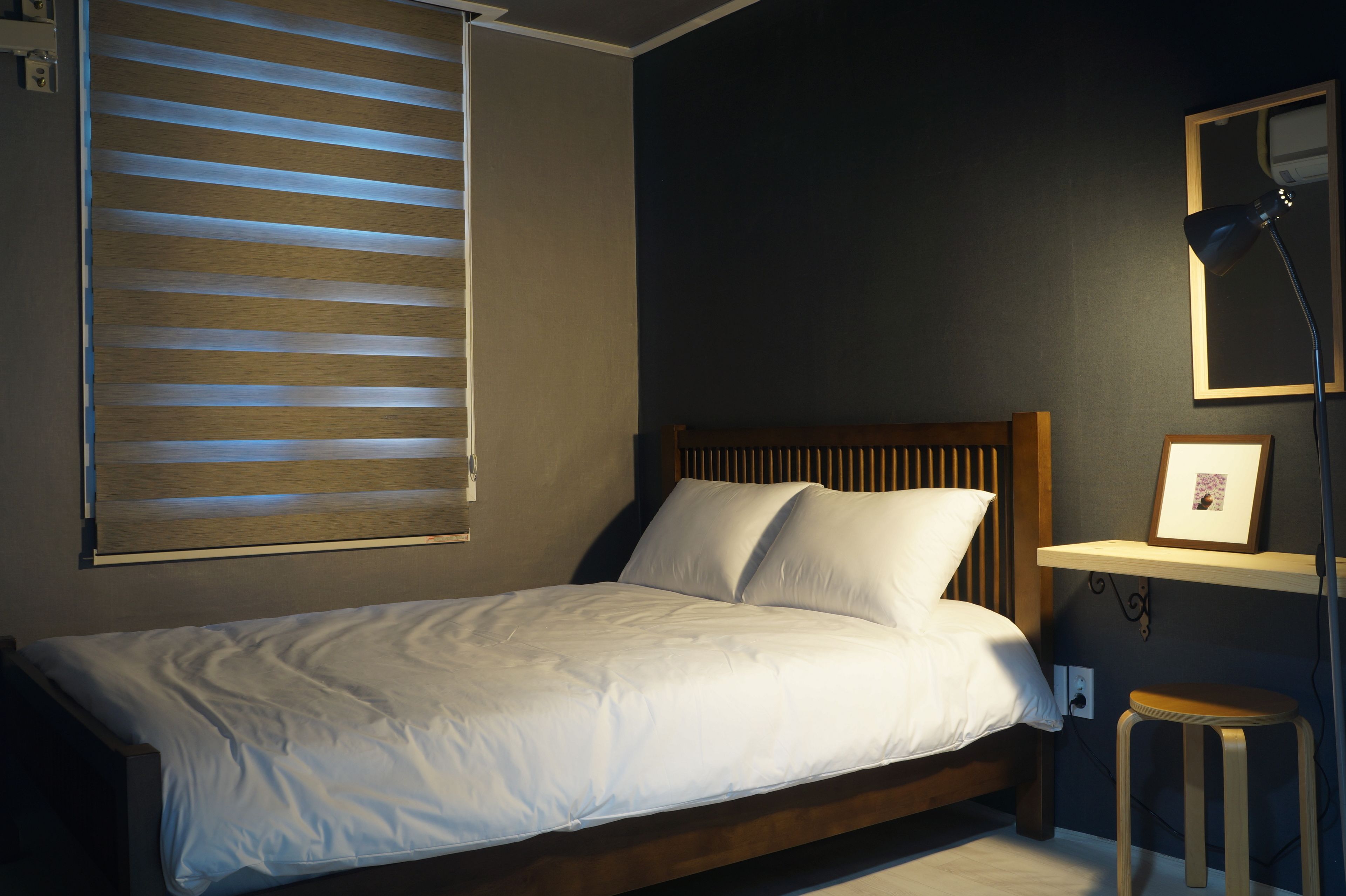 A bedroom with a bed, a nightstand, a wall-mounted shelf with a picture, and a window with blinds.