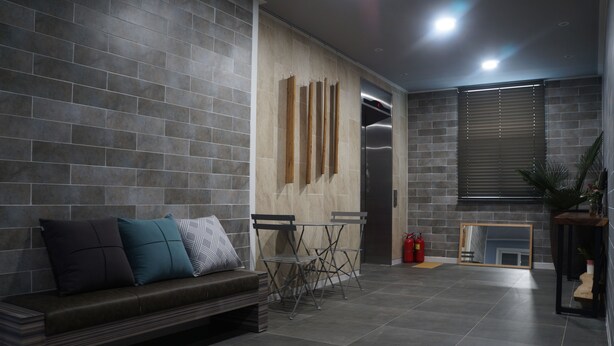 A modern lobby with a grey tiled floor, a dark brown sofa with cushions, a metal folding table, and a large mirror on the wall. - Art N Stay - Hostel (Suncheon)