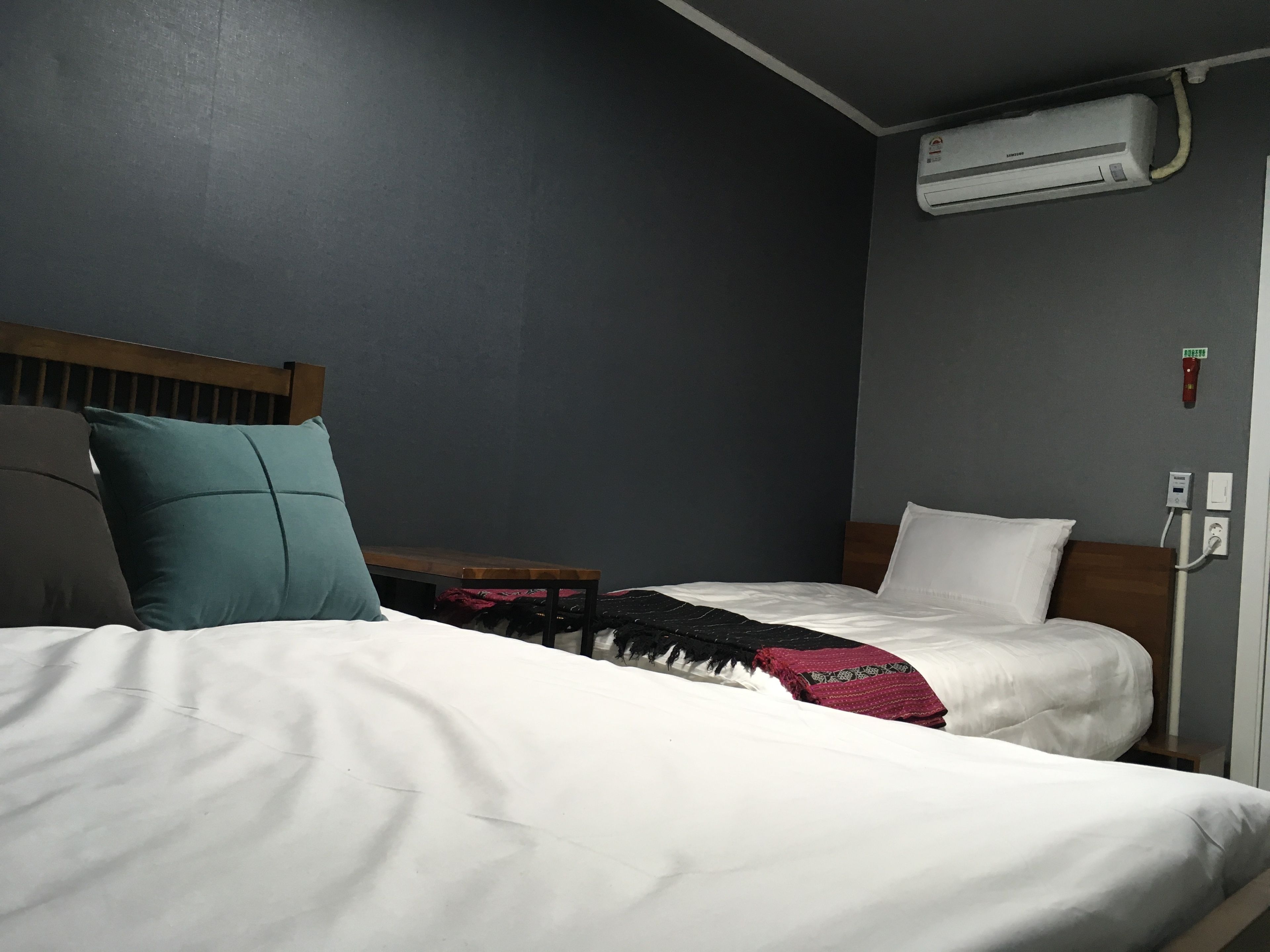 A hotel room with two beds, a wooden headboard, a wall-mounted air conditioner, and a bedside table with a red and black blanket.