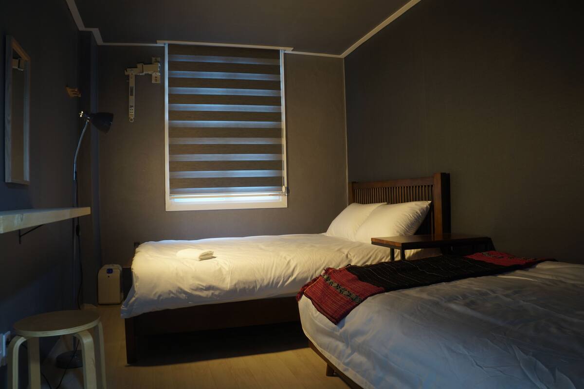 Une chambre avec deux lits, une fenêtre avec des stores, une table de chevet et un petit tabouret.