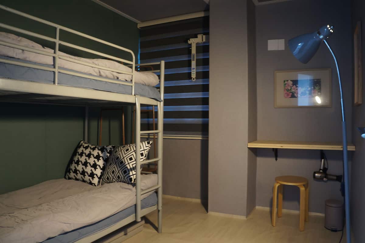 Une chambre avec un lit superposé, un bureau et un tabouret, une poubelle et un tableau encadré au mur.